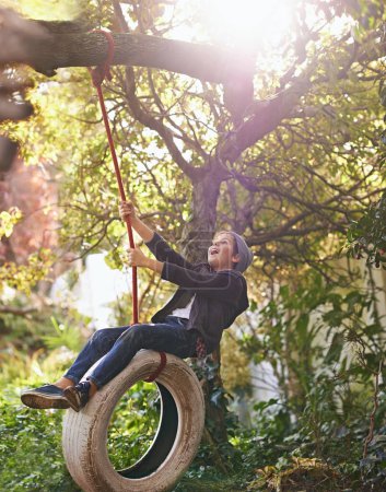 Boy, child and playing on tyre swing in garden with happiness, recreation or countryside vacation in summer. Kid, excited and diy adventure playground in backyard with sunlight and trees in nature.