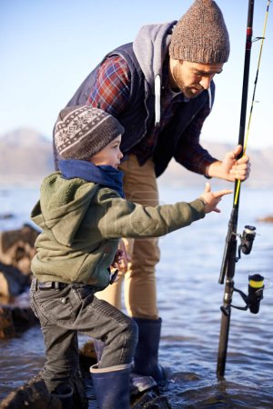 Téléchargez les photos : Père, la pêche et l'enseignement pour les jeunes garçons, les roches et les liens pour l'activité par l'océan. Mer, canne et apprendre à pêcher pour le développement de l'enfance, l'hiver et le passe-temps ensemble pendant les vacances à la mer. - en image libre de droit