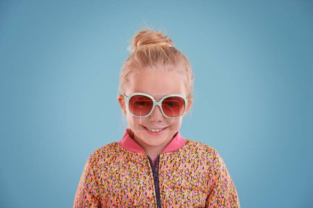 Photo for Child, portrait and fashion with sunglasses in studio for trendy, cool and funky style with confidence. Cute girl, smile and happy in blue background, young kid and retro clothes with glasses. - Royalty Free Image