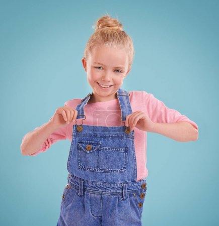 Photo for Child, portrait and smile in studio with confidence in dungarees on blue background, fashion or mockup space. Girl, school student and positive childhood in Australia with good mood, growth or kid. - Royalty Free Image