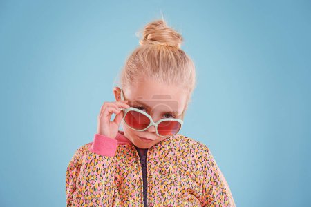 Photo for Portrait, fashion and child with sunglasses in studio with trendy, cool and funky outfit with confidence. Stylish, pout and girl kid model with cool style and accessory isolated by blue background - Royalty Free Image
