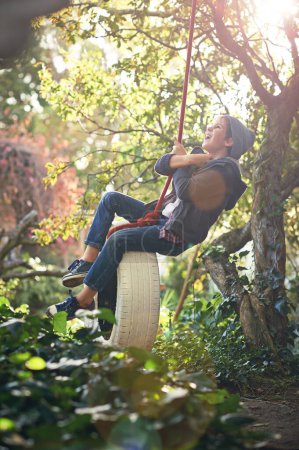 Boy, child and playing on tyre swing in garden with happiness, recreation and countryside vacation in summer. Kid, excited and diy adventure playground in backyard of home with sunlight and trees.