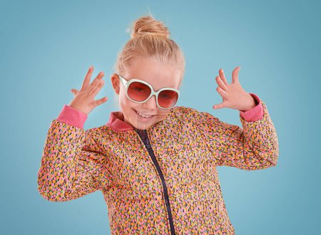 Téléchargez les photos : Petite fille, portrait et mode avec des lunettes de soleil en studio pour un style branché, cool et funky avec confiance. Enfant mignon, sourire et heureux en fond bleu, jeune enfant et vêtements rétro avec des lunettes. - en image libre de droit