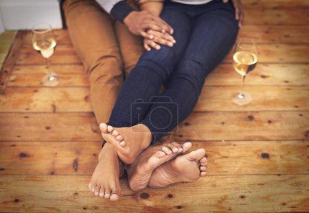 Téléchargez les photos : Vin, pieds et couple se détendre ensemble sur le sol en bois pour une date romantique à la maison moderne. Amour, champagne et gros plan des jambes d'homme et de femme avec boisson alcoolisée assis sur le sol à l'appartement - en image libre de droit