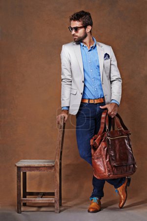 Photo for Bag, chair and confident man with clothes for fashion, style and formal wear isolated on studio background. Male person, gentleman and businessman with trendy outfit, class or suit in brown backdrop. - Royalty Free Image