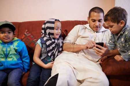 Téléchargez les photos : Enfants, père musulman et mobile dans la maison pour jouer à des jeux, bingo ou regarder des vidéos avec la technologie. Famille, papa et enfants ou garçon en portrait dans le salon pour se lier, aimer et se détendre ensemble. - en image libre de droit