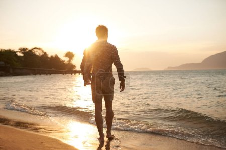 Foto de Hombre, caminando por la playa y el océano con puesta de sol, destello de lente con vista hacia atrás y olas de vacaciones en Hawai. Turismo, viajes y aventura con paz, zen y aire fresco para el bienestar, el verano y el sol. - Imagen libre de derechos