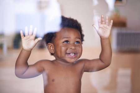 Téléchargez les photos : Bébé, heureux et sourire derrière la fenêtre, développement naturel et de l'enfant pour la croissance des tout-petits dans la maison familiale. Enfants, éducation sensorielle et toucher la main sur le verre, curieux jeu avec un jeune garçon africain. - en image libre de droit