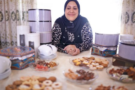 Téléchargez les photos : Femme, musulman et gâteaux dans la maison pour la célébration, table joyeuse et salle à manger avec de la nourriture pour Iftar au Ramadan. Personne islamique, dessert et joie à l'Aïd Moubarak, religion et vacances ou événement à la maison. - en image libre de droit
