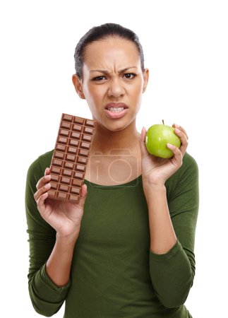 Photo for Portrait, woman and choosing chocolate versus apple for health, wellness and diet benefits. Thinking, person and choice of candy, fruits and healthy food challenge for nutritional balance in studio. - Royalty Free Image