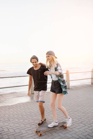 Photo for Couple, skateboard and outdoor on boardwalk, happy and helping hand for balance, learning and playful by sea. Man, woman and teaching for skating, support and fun on promenade for vacation in Italy. - Royalty Free Image