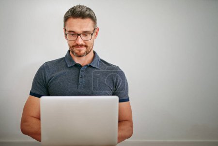 Photo for Man, wall and freelance with smile on laptop in casual for relax with social media on internet for technology. Male person, glasses and house with blog for website with typing for writer career. - Royalty Free Image