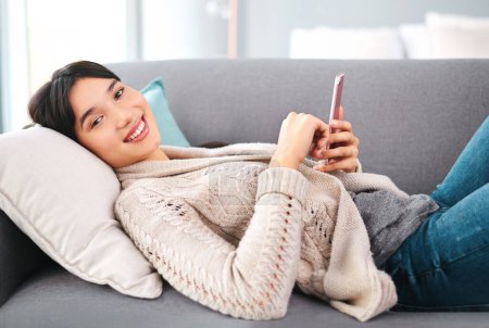 Photo for Asian woman, home and smile on sofa with smartphone, text and messages with dating app and social media. Female person, internet and online for post, content update and happy with videos in portrait. - Royalty Free Image