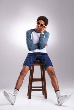 Photo for Cool guy, shades and studio chair for trendy fashion, hipster style and creative aesthetic indoor. African man or handsome fashionista and sitting on stool with confidence in clothes and sneakers. - Royalty Free Image
