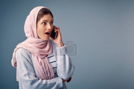 Foto de Sorprendido, llamada telefónica y mujer musulmana con teléfono inteligente en el estudio para el choque, wow u omg reacción sobre fondo azul. Hijab, sorprendido y árabe persona con expresión wtf para chismes, noticias falsas o secreto. - Imagen libre de derechos