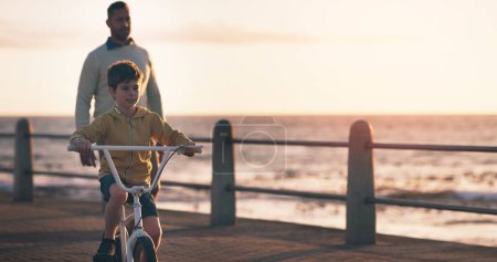 Photo for Father, boy and bike for learning at beach, promenade and sunset on vacation with care, love and bonding. Man, child and bicycle with support, teaching and outdoor by sea, waves and holiday in summer. - Royalty Free Image
