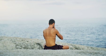 Téléchargez les photos : Calme, yoga ou homme en mer en méditation pour la paix, le bien-être ou la pleine conscience dans la nature extérieure pour se détendre. Chakra, vue holistique ou arrière du yogi sur le rocher à la plage pour la sensibilisation ou l'équilibre dans les pilates. - en image libre de droit