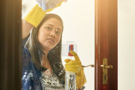 Photo for Woman, glass and cleaning door with spray in home for housekeeping chores, window or product. Female person, cloth and gloves or washing dirt as professional service for hygiene, germs or liquid soap. - Royalty Free Image