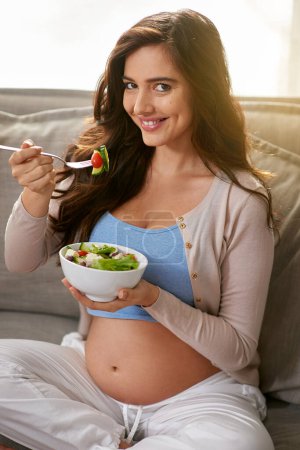 Téléchargez les photos : Sourire, manger et femme enceinte avec salade sur canapé pour le bien-être, l'alimentation et la santé à la maison. Heureuse, la maternité et la femme profitant de la nourriture, repas ou dîner avec des légumes dans le salon à la maison - en image libre de droit