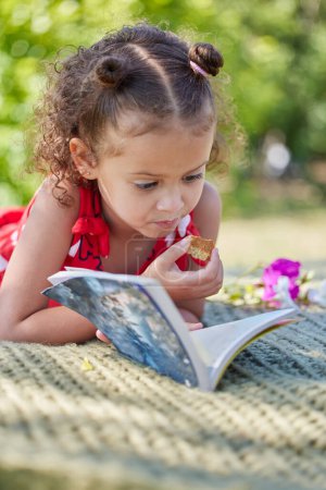 Téléchargez les photos : Fille, enfant et livre sur la couverture de pique-nique pour lire avec manger des cookies dans le parc pour se détendre avec l'apprentissage. Enfant, histoire et biscuit sur pelouse pour le développement, l'éducation et l'alphabétisation avec le soleil dans la nature. - en image libre de droit