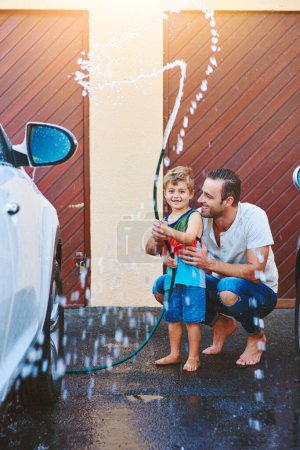 Photo for Driveway, father and child with water hose for outdoor activity or fun in summer on weekend, helping and learning. Dad, boy and spraying liquid or h2o on vehicle for washing or cleaning and splash - Royalty Free Image