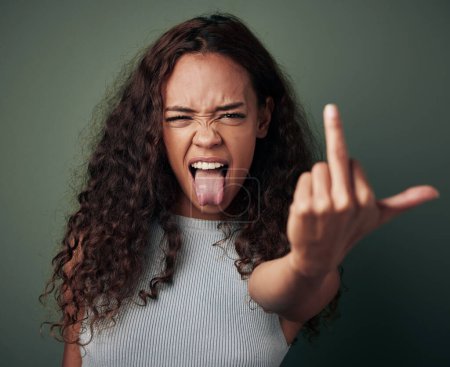 Téléchargez les photos : Portrait, woman and middle finger sign with studio for expression or conflict, rebel and fuck you for opinion. Girl, isolated and green background with hand gesture for hate with tongue out and rude - en image libre de droit