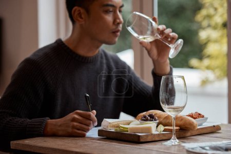 Drinking, eating and restaurant with asian man food critic writing notes for review as customer. Cheese platter, glass and wine with consumer person in eatery for gourmet cuisine or meal assessment.