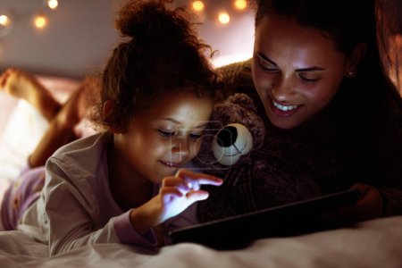 Photo for Woman, girl kid and tablet in bed, storytelling or watch cartoon at bed time with bonding and love at family home. Mother, daughter and together in bedroom at night with ebook, games or movie. - Royalty Free Image