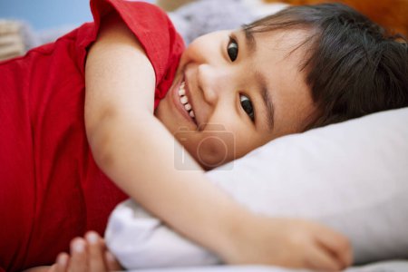 Foto de Sonríe, lindo y niño en la cama jugando y divirtiéndose en casa con actitud positiva, buena y feliz. Riendo, divertido y dulce niño asiático relajarse en la almohada para la siesta en el dormitorio en la casa moderna - Imagen libre de derechos