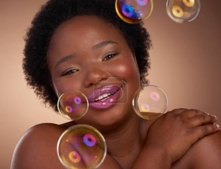 Téléchargez les photos : Joyeux, femme noire et bulles pour la beauté, les soins de la peau ou le maquillage avec brillance ou lueur dans le portrait. Modèle féminin africain et visage avec sourire, rouge à lèvres et cosmétiques pour l'auto-soin sur fond beige. - en image libre de droit