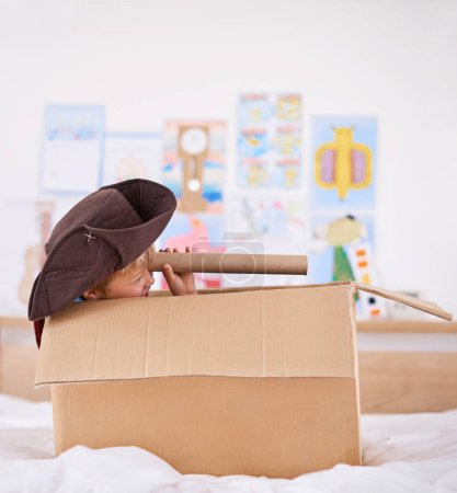 Boy, box and pirate hat with spyglasses on bed for playing, adventure and game as creativity. Kid, cardboard and home with telescope in bedroom for fun, journey and activity as imagination.
