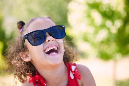 Téléchargez les photos : Fille, enfant et rire avec des lunettes de soleil dans le parc, le jardin ou la nature pour se détendre en vacances en été. Enfant, heureux et excité par la pensée, la blague et la mémoire comique au soleil avec des idées drôles en vacances. - en image libre de droit