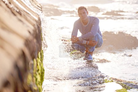 Photo for Handsome man, portrait and beach with water in relax for summer holiday, weekend or enjoying day by the sea. Young male person crouching on rocky slope by ocean waves for outdoor vacation in nature. - Royalty Free Image