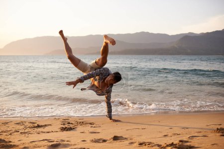 Téléchargez les photos : Homme, danseur et breakdance avec sable sur la plage pour des performances hip hop, entraînement ou portrait. Océan, danse et mouvement avec énergie par horizon, cascade ou talent flexible pendant les vacances aux Maldives. - en image libre de droit