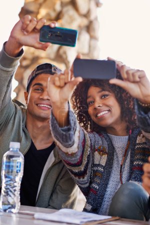 Foto de Estudiantes, amigos y selfie al aire libre en el campus para publicar en línea redes sociales, conexión o vinculación. Hombre, mujer y sonrisa en la mesa de almuerzo para comunicarse juntos con la tecnología, internet o relajarse. - Imagen libre de derechos