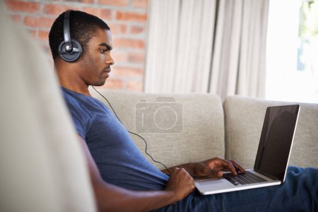 Foto de Hombre, internet y música con portátil en el sofá para la transmisión de películas, podcast y juegos en línea con escucha. Joven, negro y tecnología con auriculares para relajarse, estudiar y aprender en el salón. - Imagen libre de derechos