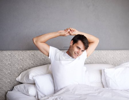 Photo for Man, home and stretch in bed, adult and smile for daily routine and rest and wellness. Male person, happy and bedroom in sleepwear and morning for waking up and relaxing on Saturday day off - Royalty Free Image