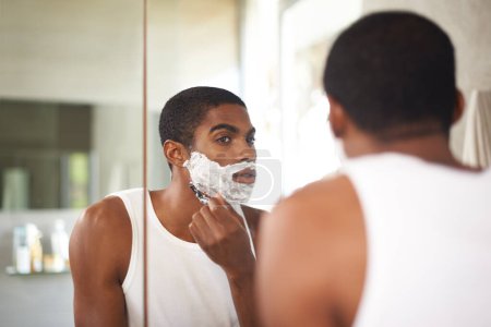 Photo for Black man, mirror and shaving with razor in bathroom for grooming, skincare or morning routine. Reflection, beard or person with cream on face for cleaning, health or hair removal for hygiene in home. - Royalty Free Image
