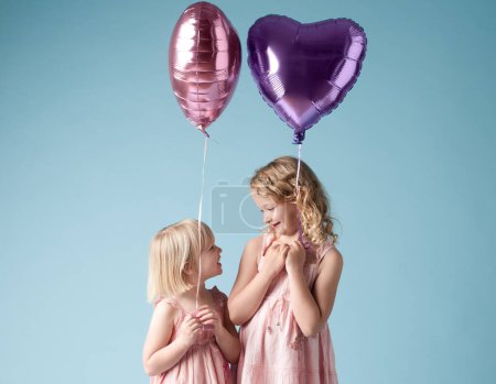 Foto de Children, sister and smile with heart balloon in studio on blue background for family or party. Birthday, bonding or celebration with girl kid friends, happy on color backdrop together for event. - Imagen libre de derechos