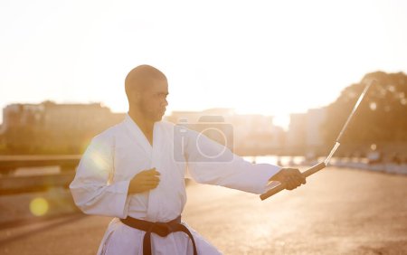 Photo for Man, karate and fitness with nunchuck in city for fighting style, technique or combat skill. Young male person or taekwondo fighter with weapon in martial arts or self defense on street in urban town. - Royalty Free Image
