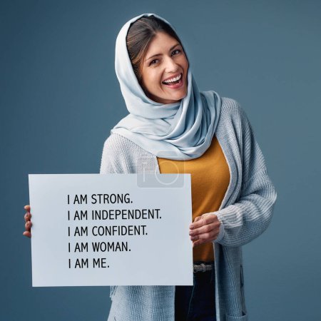 Muslima, Frau und glücklich mit Plakat im Atelier auf blauem Hintergrund für Gleichberechtigung, Empowerment und Aktivistin. Weibliche Person, Karte und Lächeln vertrauensvoll für Menschenrechtskampagne und Fürsprecherin im Porträt.