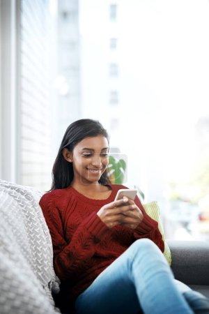 Photo for Relax, smile and Indian woman on couch with phone for social media, networking or reading on weekend. Happy, scroll and girl on sofa with smartphone for communication, online chat or search in home. - Royalty Free Image