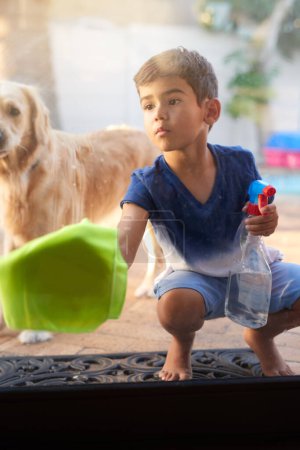 Foto de Rocíe el frasco, el paño y la ventana de limpieza infantil para evitar la suciedad, los gérmenes o las bacterias con el perro en casa. Detergente, higiene y niño limpian la puerta de vidrio para desinfectar con cachorro de mascota en el patio trasero de la casa - Imagen libre de derechos