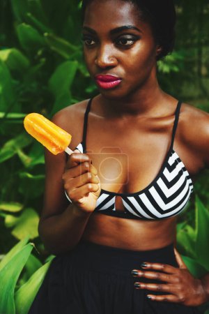 Téléchargez les photos : Femme noire, mode et style avec popsicle dans le jardin avec confiance pour glamour, soutien-gorge et tenue d'été. Femme, vêtements et look tendance en plein air sur le temps chaud pour énervé et moderne - en image libre de droit