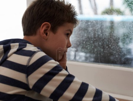 Photo for Window, rain and sad boy child on a floor unhappy, upset or annoyed by bad weather in his home. Glass, depression and kid in a living room frustrated by storm, cold or unexpected winter disaster. - Royalty Free Image
