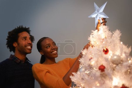 Photo for Couple, Christmas and tree with star decoration for holiday celebration in winter season for festive, giving or presents. Man, woman and lights in living room for bonding vacation, event or tinsel. - Royalty Free Image