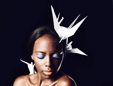 Téléchargez les photos : Femme noire, maquillage et beaux-arts avec des cosmétiques, des oiseaux origami ou colombe sur fond de studio sombre. Gros plan ou visage d'une femme africaine ou d'un mannequin avec une beauté ou une ombre à paupières bleue sur un espace maquillé. - en image libre de droit
