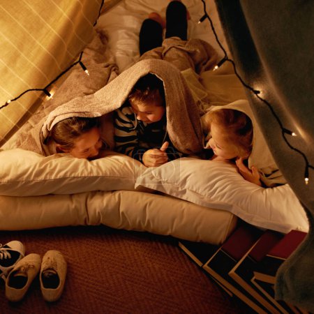 Photo for Children, happy and sleepover in tent at night with conversation, bonding and holiday adventure or vacation. Young friends or kids by fairy lights, pillows and blanket at home for fun storytelling. - Royalty Free Image