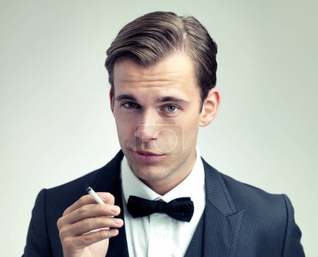 Retrato, hombre elegante o joven fumando un cigarrillo en estudio, formal o de moda vintage por fondo blanco. Cara, actitud o caballero en traje retro o esmoquin, pajarita o hábito de tabaco en evento.