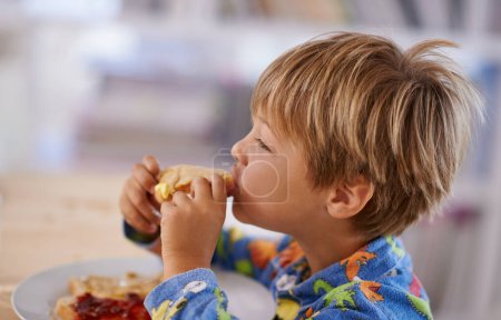 Photo for Food, breakfast and boy eating a sandwich in morning, pyjamas and home for nutrition. Children, hungry and meal for young male for lunch, childhood development and bread for health and wellness. - Royalty Free Image